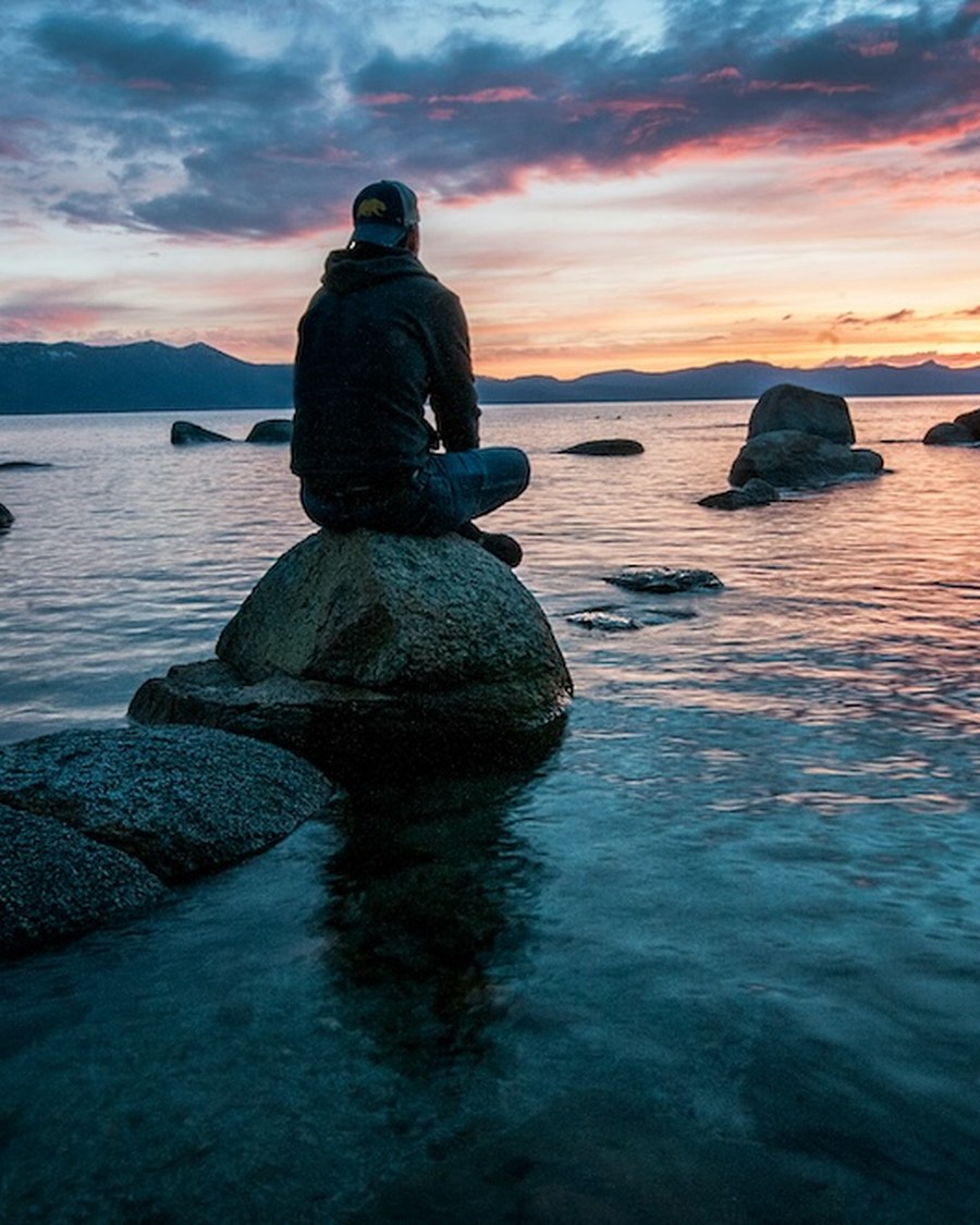 A person sitting peacefully on a rock by the water at sunset, reflecting